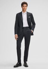 Donkergrijs op maat gemaakt pak met een slim fit, voorzien van een enkelvoudig jasje, revers, en bijpassende broek. Witte overhemden eronder.