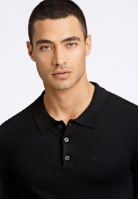 Svart polo-skjorte laget av ribbestrikket stoff, med klassisk krage og tre-knapps plaske, med subtil logo detaljer på brystet.