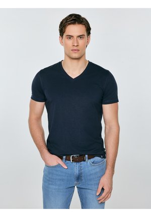 Młody mężczyzna z krótkimi brązowymi włosami, ubrany w czarny T-shirt z dekoltem w serek i jasnoniebieskie dżinsy, stojący z jedną ręką w kieszeni na jednolitym tle.