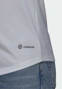 Camiseta deportiva blanca con una textura suave, un sutil patrón geométrico y el logo "AEROREADY" cerca del dobladillo, combinada con tejido de mezclilla.