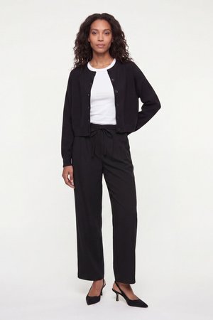 Femme aux cheveux bouclés portant un cardigan noir boutonné, un t-shirt blanc, un pantalon noir à cordon de serrage et des talons slingback noirs sur fond blanc.