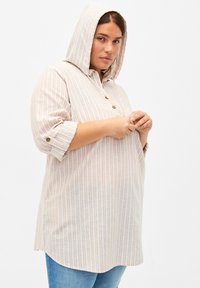 Kapuzená tunika světlé béžové barvy s vertikálními pruhy, předním zapínáním na knoflíky a ohrnutými rukávy, nabízející volný střih a měkkou texturu.