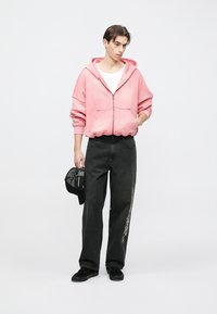 Rosa Kapuzenpullover zum Reißverschluss mit lockerer Passform, kombiniert mit hoch taillierten schwarzen Jeans mit seitlicher Stickerei. Schwarze Schuhe und Kappe vervollständigen den Look.