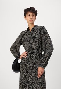 Black paisley-patterned dress with long sleeves, a shirt collar, and a tied waist. Fabric shows a textured design with gold accents.