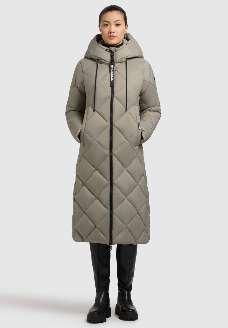 Cappotto lungo trapuntato color verde oliva con cappuccio, chiusura con zip e cordino. Presenta cuciture diagonali e due tasche laterali. Abbastanza abbinato a stivali neri.