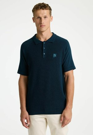 Mann trägt ein dunkelblaues Polohemd mit kurzen Ärmeln und drei Knöpfen sowie cremefarbene Hose, steht vor einem schlichten weißen Hintergrund.