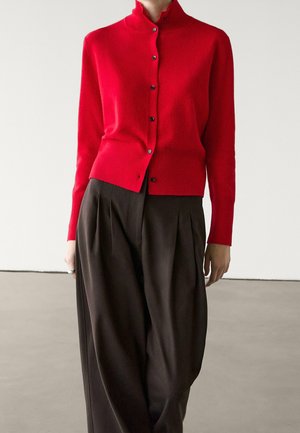 Roter Cardigan mit Knöpfen und hohem Kragen, kombiniert mit lockeren, dunkel plissierten Hosen, getragen von einer stehenden Person vor einem schlichten Hintergrund.