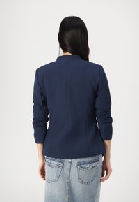 Námořnický modrý přiléhavý blazer s vysokým výstřihem, dlouhými rukávy a nabíranými manžetami, kombinovaný se světle modrou džínovou sukní s zadními kapsami.