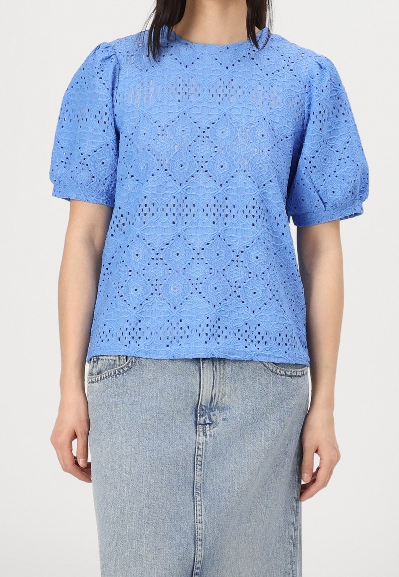 Femme portant un chemisier en dentelle bleu clair à manches courtes bouffantes et une jupe en jean clair, debout devant un fond uni.
