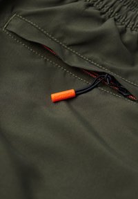 Dunkelgrüner Stoff mit einer geöffneten Tasche, schwarzem Reißverschluss und einem orangefarbenen Zuganhänger mit der Aufschrift "SUPERDRY" an einer schwarzen Schnur.