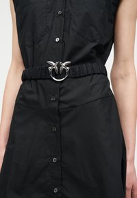 Μαύρο φόρεμα με κουμπιά και δύο τσέπες στο στήθος, με ζωνάκι ελαστικό στη μέση που διαθέτει ασημένια αγκράφα σε σχήμα δύο πουλιών που κοιτάζονται το ένα το άλλο.