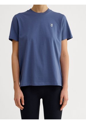 Personne portant un t-shirt bleu uni à manches courtes avec un petit logo chevron blanc sur la poitrine gauche et un pantalon noir côtelé.