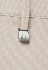 Beżowy materiał z bliskim ujęciem metalowego suwaka z wyrytym kątowym L i symbolem strzały.