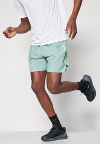 Ανοιχτοπράσινα αθλητικά σορτς με αθλητικό σχέδιο και διχτυωτές λεπτομέρειες, συνδυασμένα με μαύρα αθλητικά παπούτσια και μαύρες κάλτσες μέχρι τον αστράγαλο.