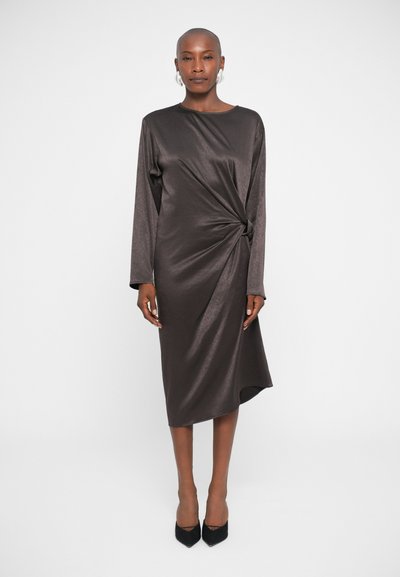 Satin, dunkelbraunes Kleid mit langen Ärmeln und einem gedrehten Frontdetail. Knielanger Schnitt mit einem dezenten Glanz, kombiniert mit schwarzen High Heels.