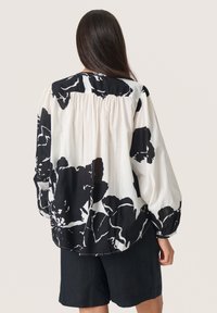 Blumenbluse in Schwarz und Weiß, mit lockerer Passform, gerafftem Rückendetail und Puffärmeln. Kombiniert mit dunklen Shorts.