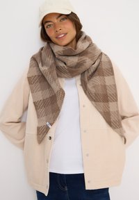 Donna che indossa un berretto beige, una grande sciarpa a quadri marrone testurizzata, una giacca di corduroy crema, una camicia bianca e jeans blu scuro su sfondo bianco.