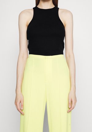 Kobieta ubrana w bezrękawnikowy czarny prążkowany top zestawiony z wysokim stanem i szerokimi, jasnżółtymi spodniami, stojąca na jednolitym tle.