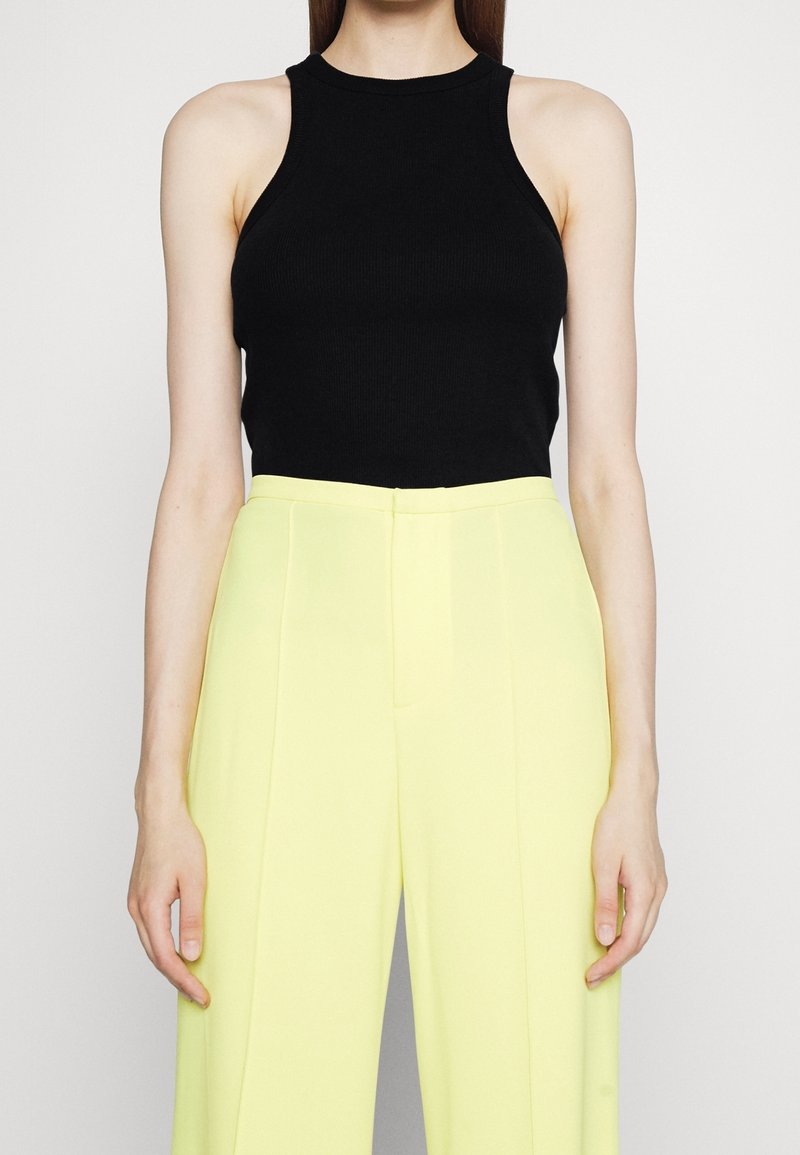 Femme portant un haut noir côtelé sans manches associé à un pantalon large taille haute jaune clair, debout devant un fond uni.