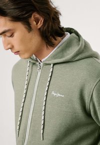 Joven con cabello castaño que lleva una sudadera verde claro con cierre y cordones blancos y negros, con el logo de Pepe Jeans en el pecho.