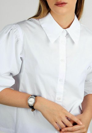 Camicia bianca con colletto e maniche a sbuffo, con bottoni sulla parte anteriore. Orologio da polso nero con quadrante argentato e cinturino in pelle su una mano.