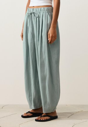 Kobieta ubrana w luźne, jasnozielone spodnie ze sznurkiem w pasie i bocznymi kieszeniami oraz czarne płaskie sandały, stojąca na kafelkowanej podłodze.