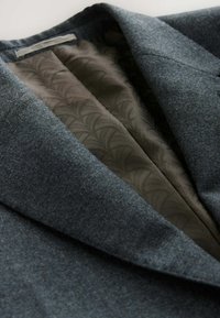 Grau wOL-blazer mit Reverskragen, mit einem gemusterten dunkelgrauen Innenfutter sichtbar. Glatte Textur und maßgeschneiderte Designdetails.