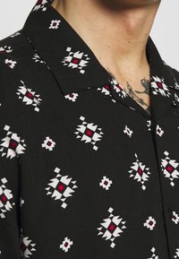 Černé tričko s krátkým rukávem s geometrickými bílými a červenými vzory. Límec je plochý a látka vypadá hladce a lehce.