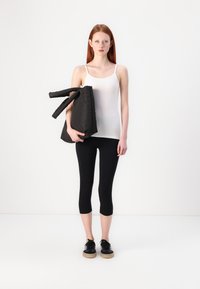 Hvit tanktopp, svarte cropped tights og svarte joggesko; modellen holder en svart polstret veske med to håndtak; glatte teksturer, minimalistisk design.