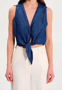 Haut en denim sans manches avec un décolleté en V profond et un devant noué, présentant une texture légère et un design structuré. Porté avec un pantalon beige.