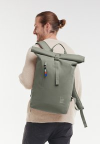 Mochila verde de tipo roll-top hecha de materiales reciclados. Cuenta con un bolsillo delantero con cremallera, correas ajustables y una textura suave con una marca mínima.