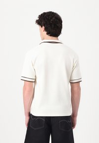 Vit kortärmad polo med ribbad krage och bruna accentlinjer. Texturerad tyg med avslappnad passform och rak fåll.