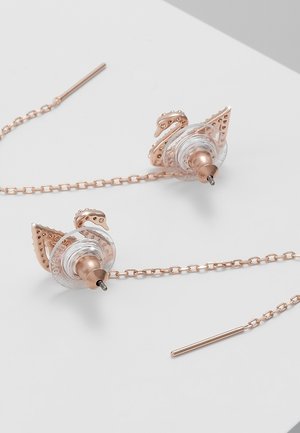 Boucles d'oreilles fil en forme de cygne en or rose avec des supports en silicone transparent placées sur une surface blanche.