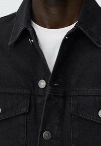 Giacca di jeans nera con colletto a punta, cuciture bianche, accessori in argento e due tasche anteriori. Indossata sopra una maglietta bianca.