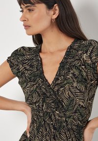 Schwarzes Kleid mit einem blättrigen grünen und beige-blumigen Muster, kurzen Flügelärmeln, tiefem V-Ausschnitt und elastischer Taille für eine figurbetonte Silhouette.