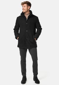 Abrigo de mezcla de lana negro con cuello alto, cierre de botones y bolsillos laterales, combinado con pantalones grises y botas negras hasta el tobillo.