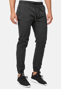 Homme portant un pantalon de jogging noir avec des poignets élastiques et une taille à cordon, associé à des baskets Adidas noires et blanches.