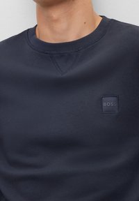 Marinblå crewneck sweatshirt gjord av slät tyg. Har en liten fyrkantig logotyp i mörkblått på bröstet och subtila sömdetaljer.
