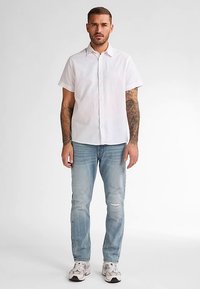 Camicia bianca a maniche corte con bottoni, jeans azzurri slavati e sneaker grigie. Braccia tatuate visibili, in piedi contro uno sfondo semplice.