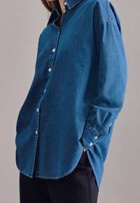 Camicia blu in denim con colletto classico, maniche lunghe e bottoni ai polsini. Presenta una vestibilità morbida e orlo curvo.