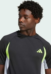 Černé sportovní tričko s krátkým rukávem, se zelenými a šedými akcenty, kulatým výstřihem a výrazným logem se třemi pruhy na hrudi.