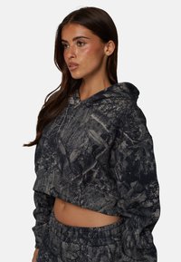 Krótka bluza z kapturem w ciemnoszarym kolorze z wzorem przypominającym korę drzewa. Posiada kaptur, długie rękawy i teksturowany materiał. Dobrane dresy dopełniają całość.