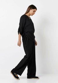 Black linen top with a V-neck and side ruching; paired with loose black trousers and espadrille shoes featuring a flat sole and strap.
