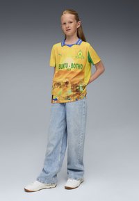 Camiseta de fútbol amarilla con detalles verdes, con un diseño gráfico y el texto "BUNTU-BOTHO". Se lleva con jeans de color azul claro y zapatillas blancas.