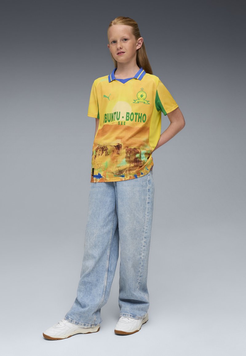 Camiseta de fútbol amarilla con detalles verdes, con un diseño gráfico y el texto "BUNTU-BOTHO". Se lleva con jeans de color azul claro y zapatillas blancas.