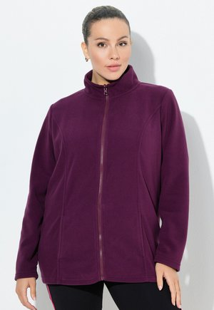 Woman wearing a zip-up purple fleece jacket with high collar, standing against a white wall with hands relaxed by sides.