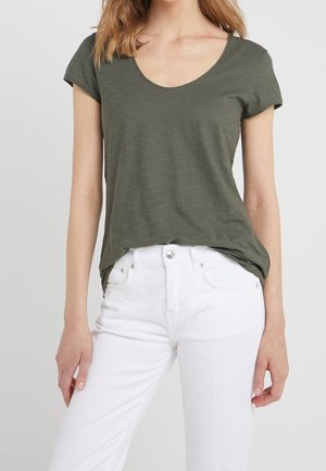 Vrouw draagt een olijfgroene, korte mouwscoopnek T-shirt, ingestopt in een hoogtailleren witte spijkerbroek, staand tegen een effen achtergrond.