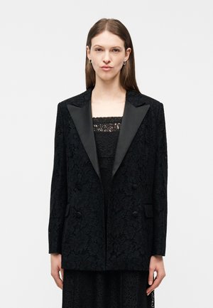 Femme portant un blazer en dentelle noire avec des revers en satin, par-dessus un top en dentelle noire assorti, debout devant un fond clair uni.