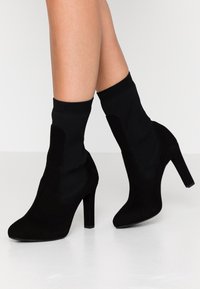 Botas negras hasta el tobillo con un tacón alto, confeccionadas en suave ante y con una parte superior de estilo calcetín tejido para mayor comodidad y estilo.