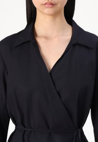Black wrap dress with a collared neckline and long sleeves, featuring a tie at the waist and a smooth, matte texture.